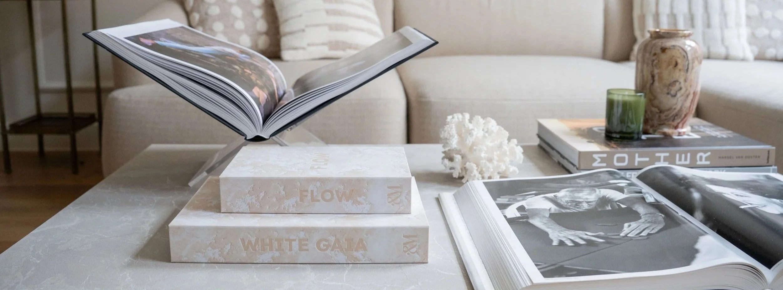 Een tafel met opgestapelde opbergboeken, een open fotoalbum, een tak van koralen, en twee gekleurde kaarsen, met een bank op de achtergrond.