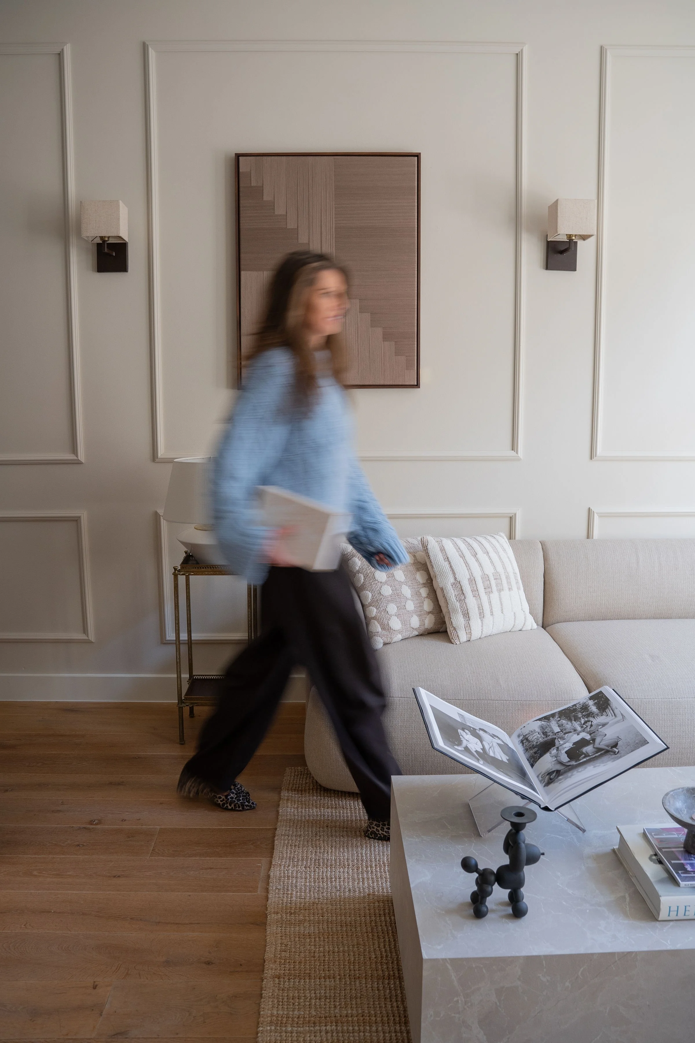 Een vrouw draagt een laptop en loopt door een woonkamer met een bank, kussens en een salontafel met foto's en boeken.