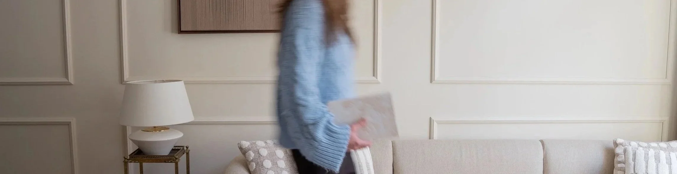 Een persoon loopt in een woonkamer, met een licht gekleurd bank en een witte lamp op een tafel.