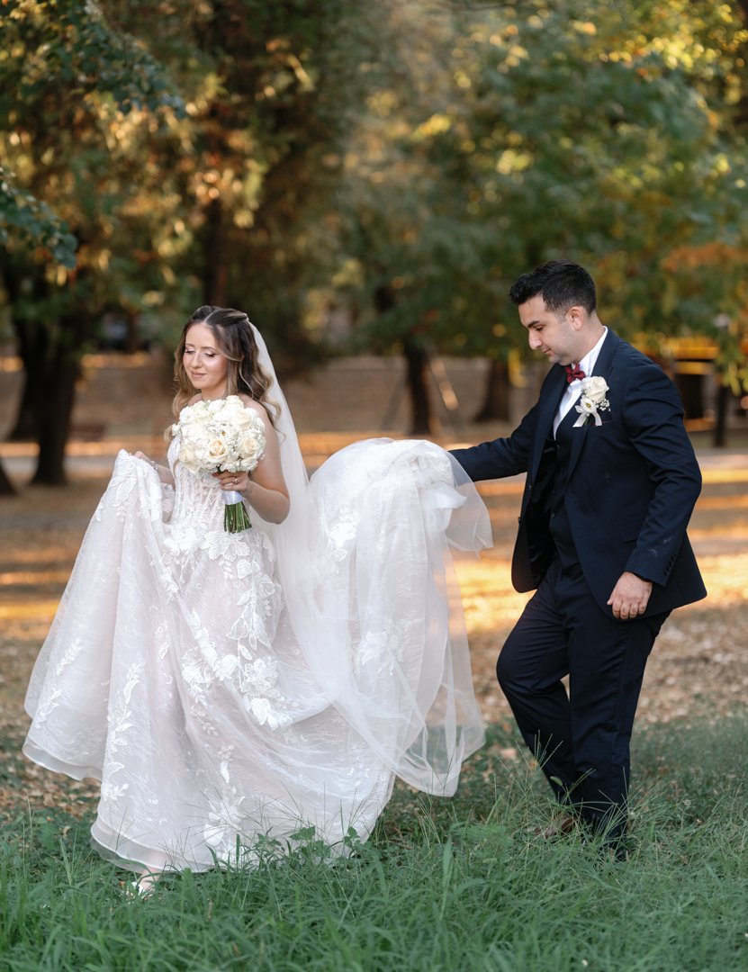 A bride wearing a wedding gown holding a bouquet of white roses and a groom in a dark suit with a boutonniere, outdoors in a park during daytime.