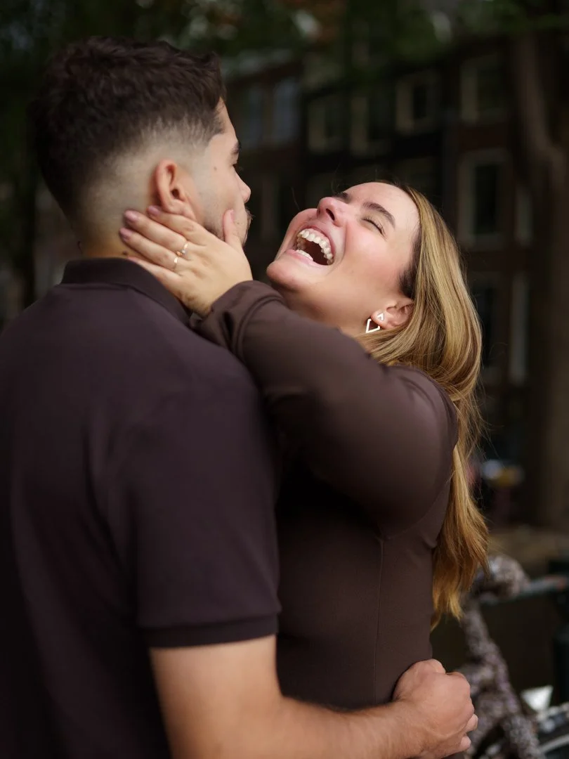 A couple is hugging and laughing outdoors, with the woman looking up at the man and both appearing happy.