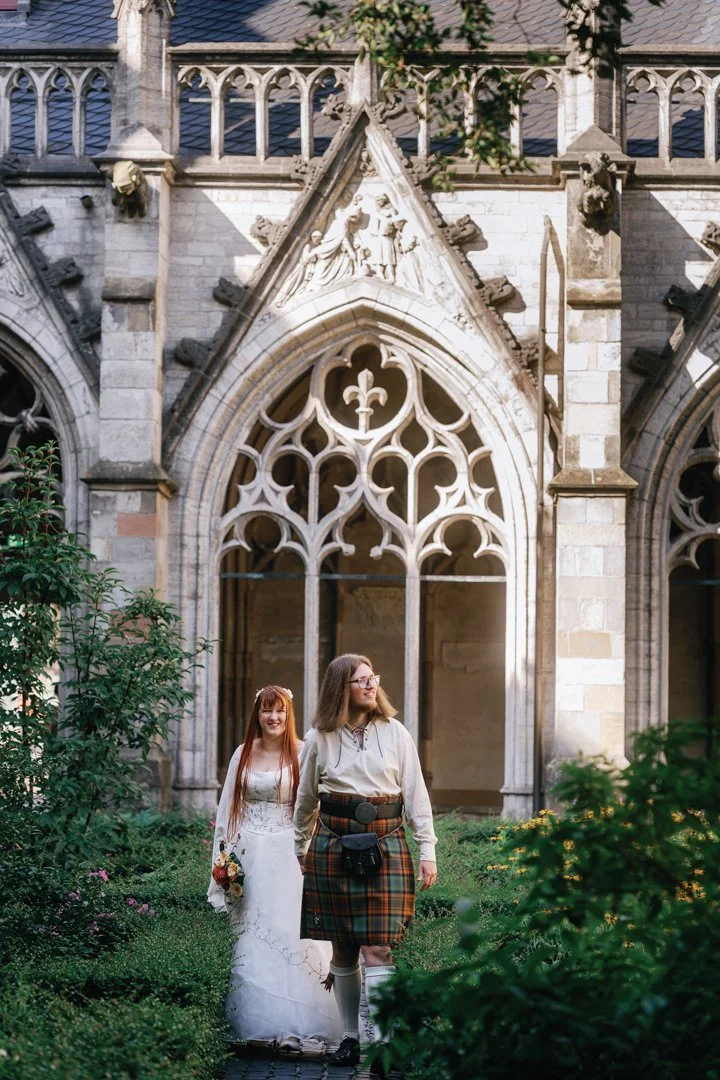 A bride and a woman walking outside a Gothic-style church with intricate stained glass windows and stone architecture.
