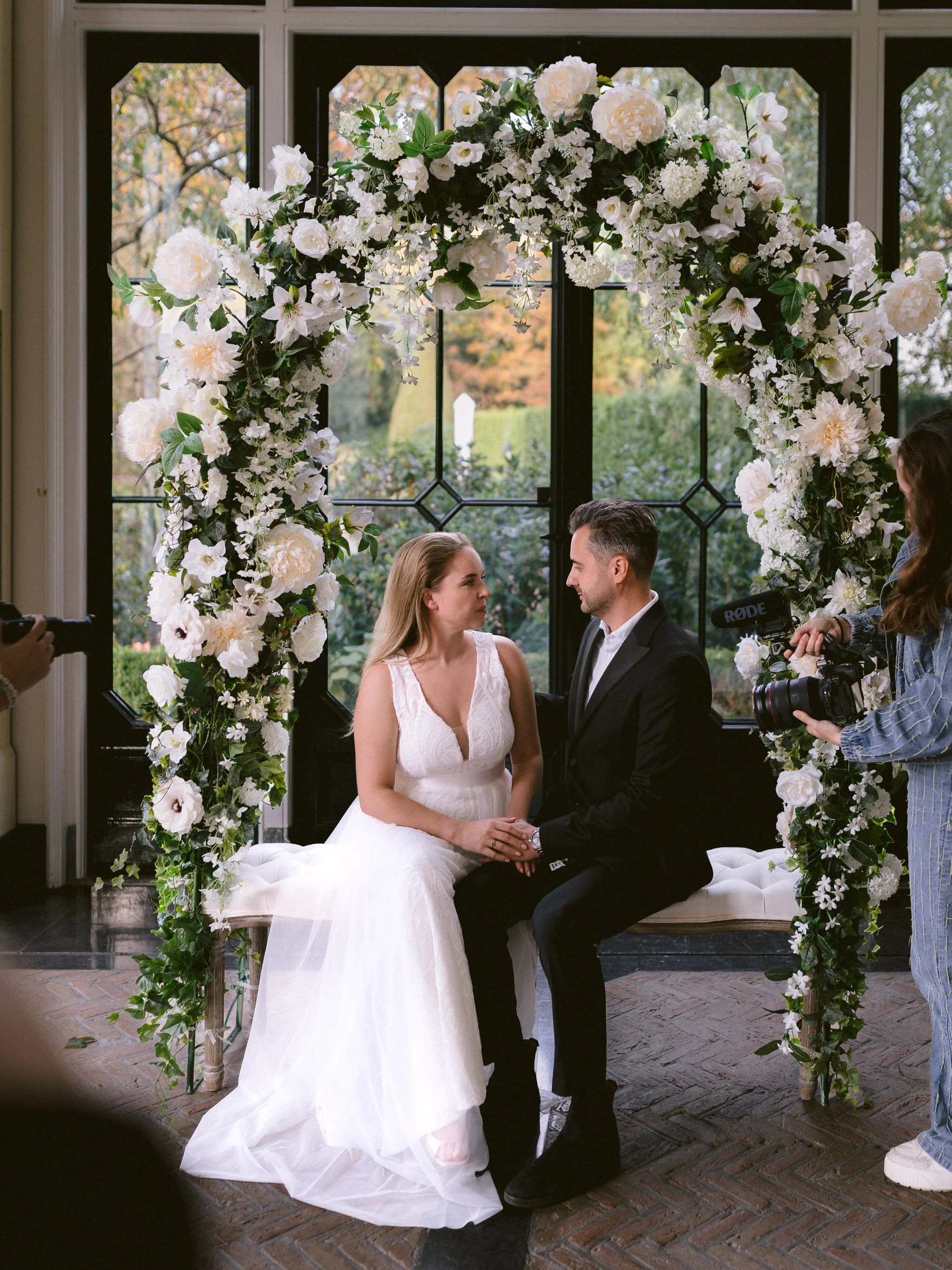 A bride and groom sitting face to face during their wedding ceremony under a floral arch with white flowers and green leaves, inside a building with large windows showing a garden outside. A camera crew is recording the event.