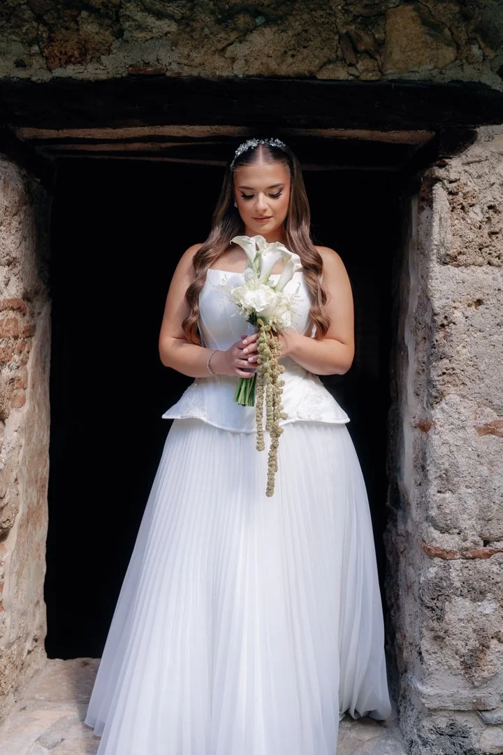 A bride in a white wedding dress holding a bouquet of white flowers, standing in a stone archway.