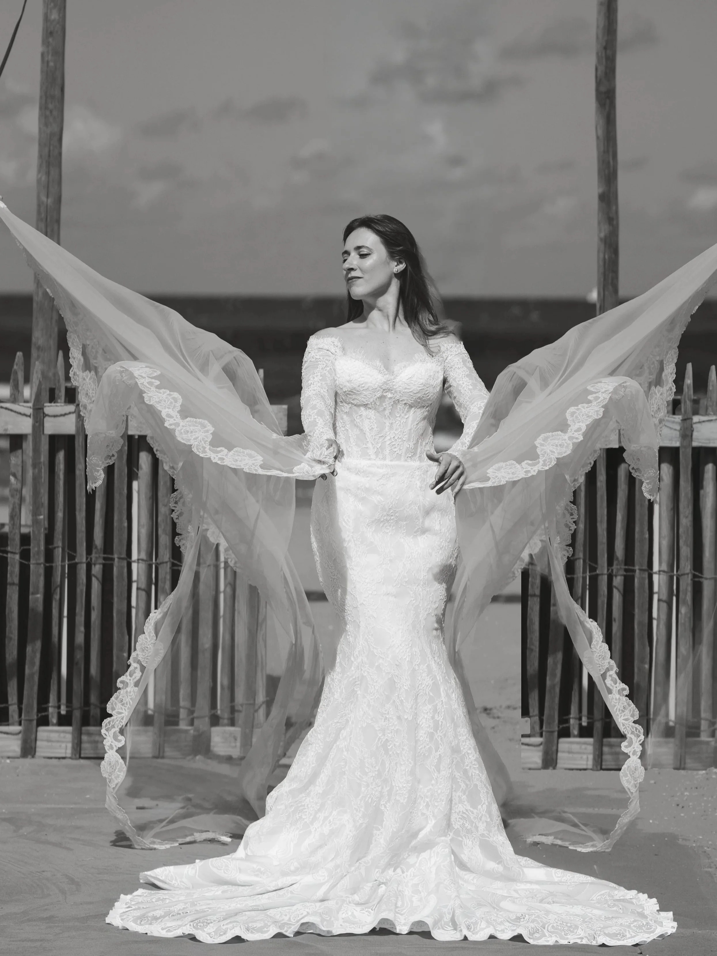 A bride in a wedding dress stands on a beach, holding the flowing lace train of her dress, with a wooden fence and cloudy sky in the background.