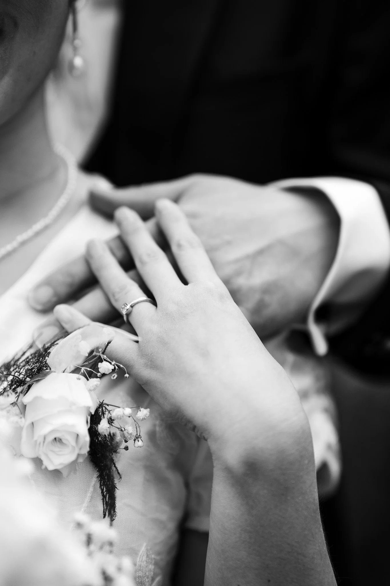 Close-up of a bride's hand with rings resting on her chest, holding a bouquet of flowers, with part of her wedding dress visible in black and white.