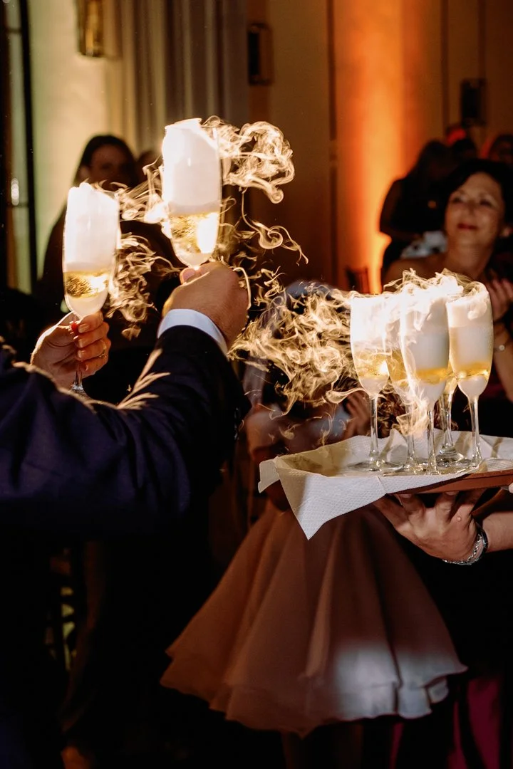 People celebrating with champagne glasses, some glasses have sparklers inside, creating a smoky effect at a festive event.