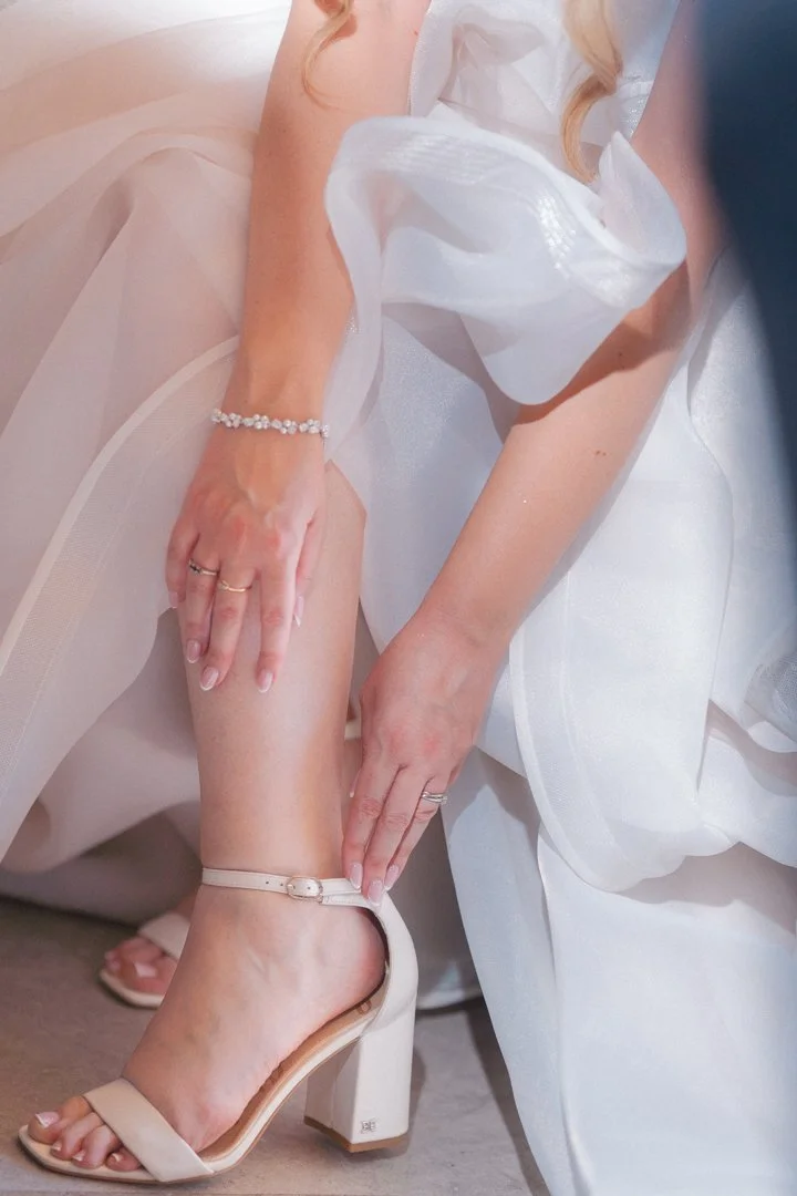 A bride in a white wedding dress adjusting her ankle strap on her high heel sandal.