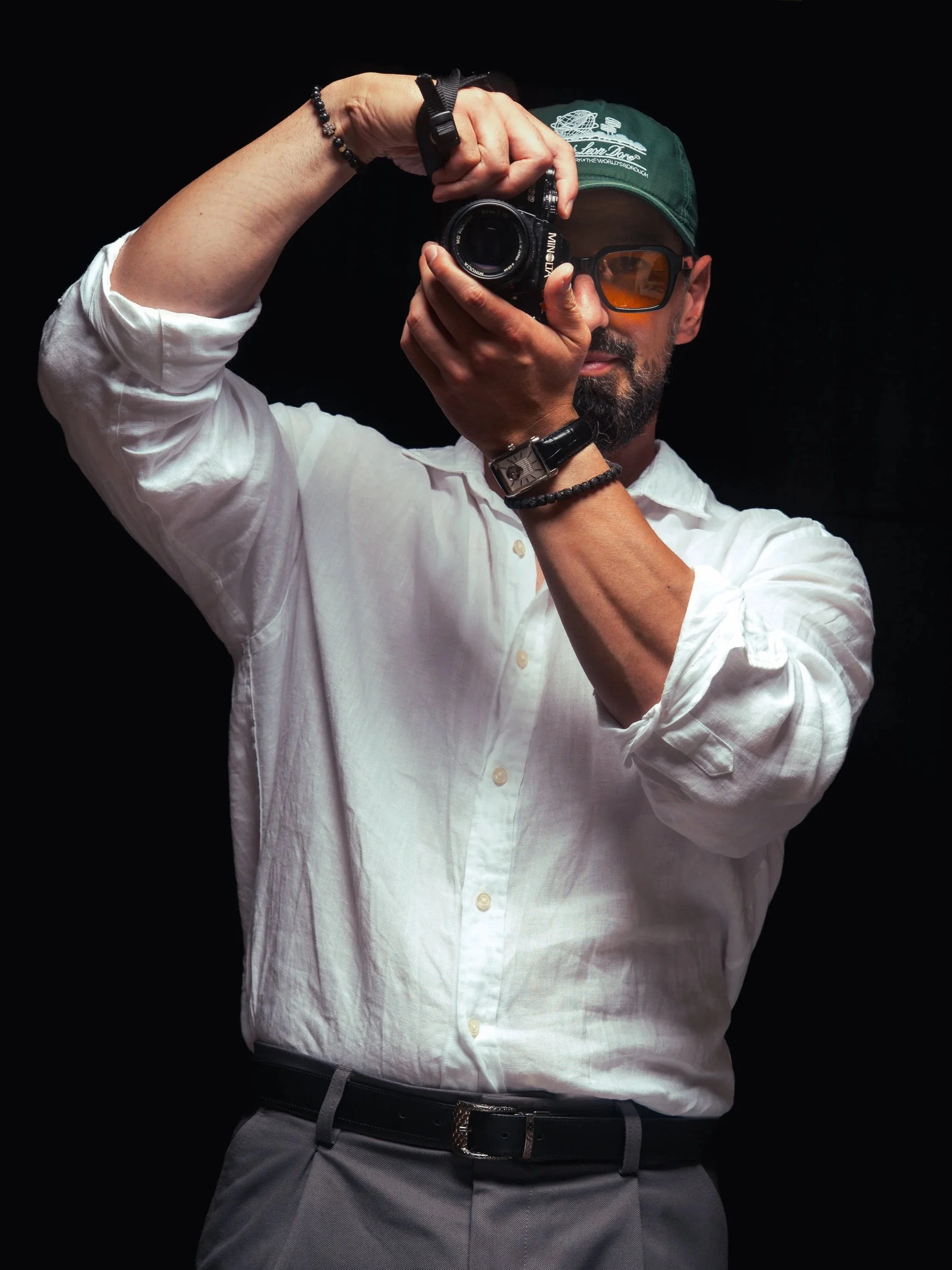 Man taking a photo with a camera, wearing sunglasses, a green cap, a white shirt, and gray pants, against a black background.