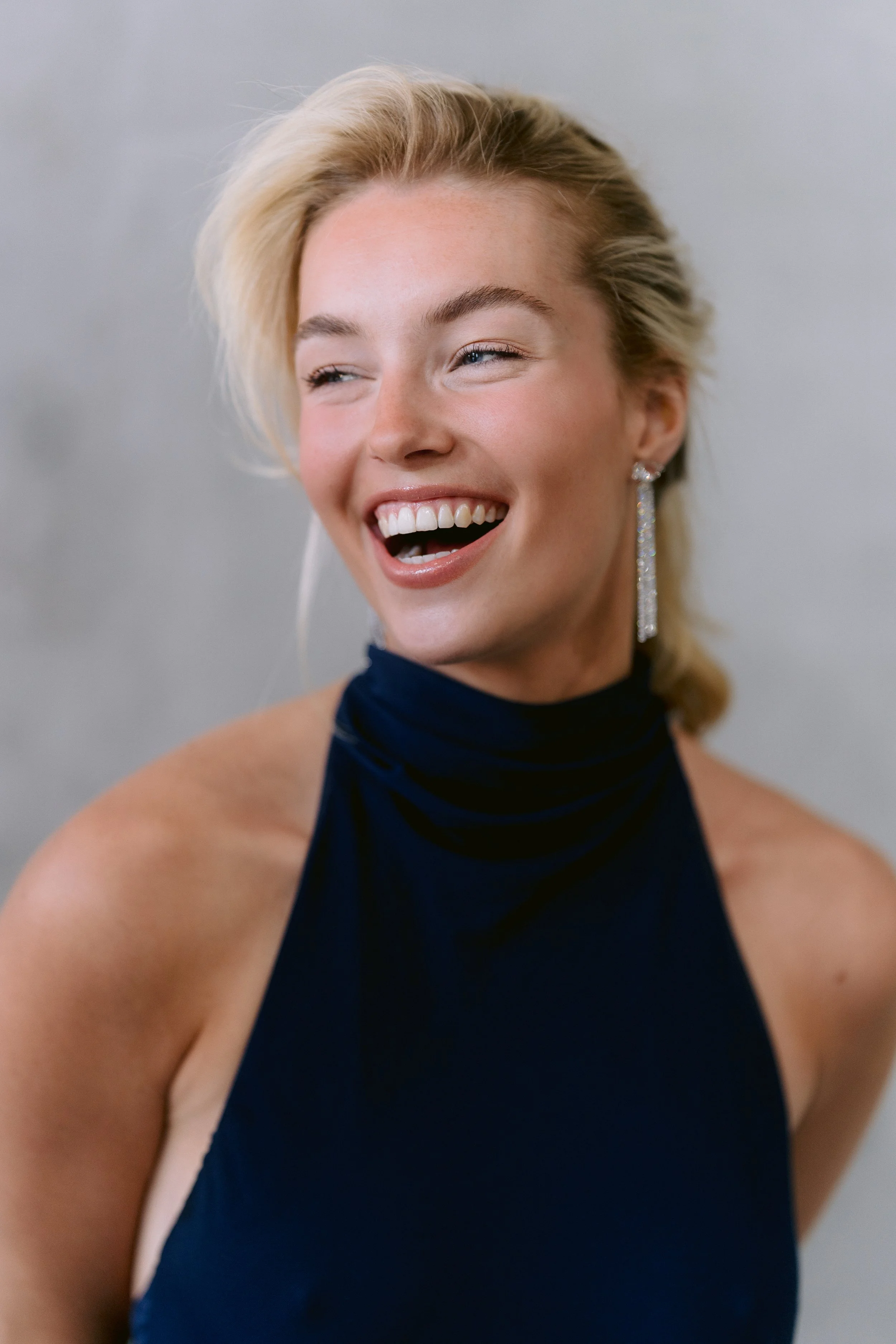 Close-up of a woman with blonde hair, laughing with her mouth open, wearing a blue sleeveless top and long dangling earrings, against a plain light gray background.