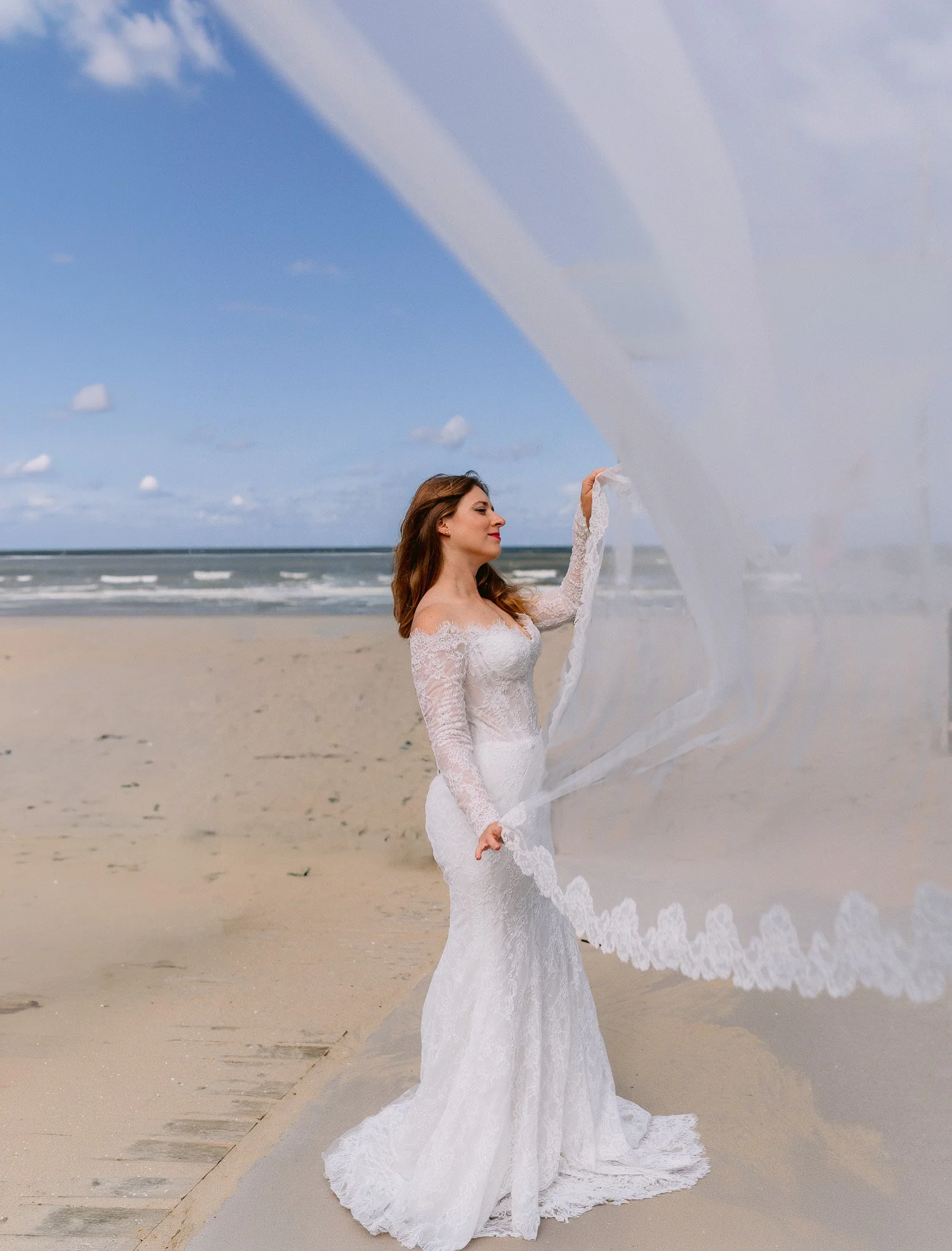 A woman in a white lace wedding dress stands on the beach, holding a flowing veil in front of her with a calm seascape and blue sky background.