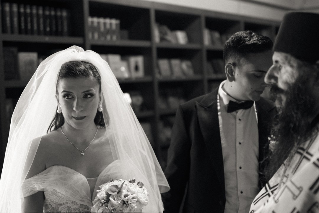 A bride in a wedding gown and veil holding a bouquet stands near a groom and a religious figure during a wedding ceremony.