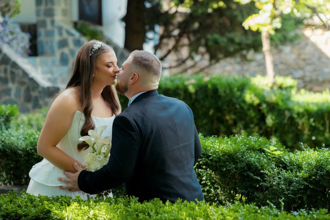 A bride and groom share a kiss outdoors surrounded by greenery and bushes.