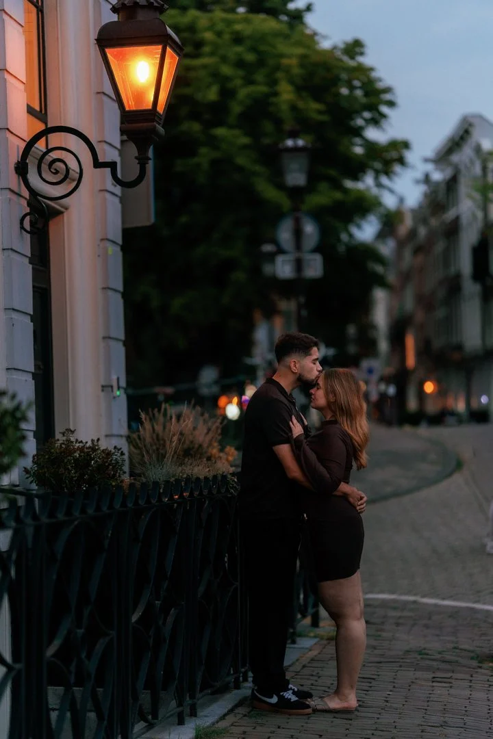A couple stands close together on a street at dusk, sharing an intimate moment near a lit street lamp. The man is gently holding the woman as they look into each other's eyes.
