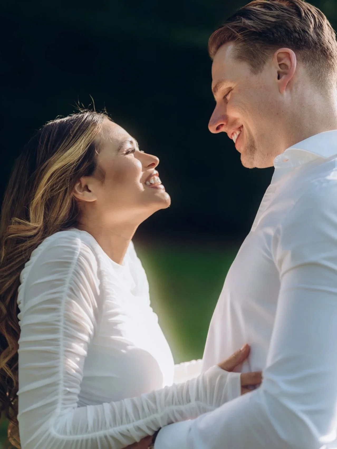 A smiling couple enjoying a moment together outdoors on a sunny day, facing each other closely, with the woman wearing a white wedding dress and the man in a white shirt.