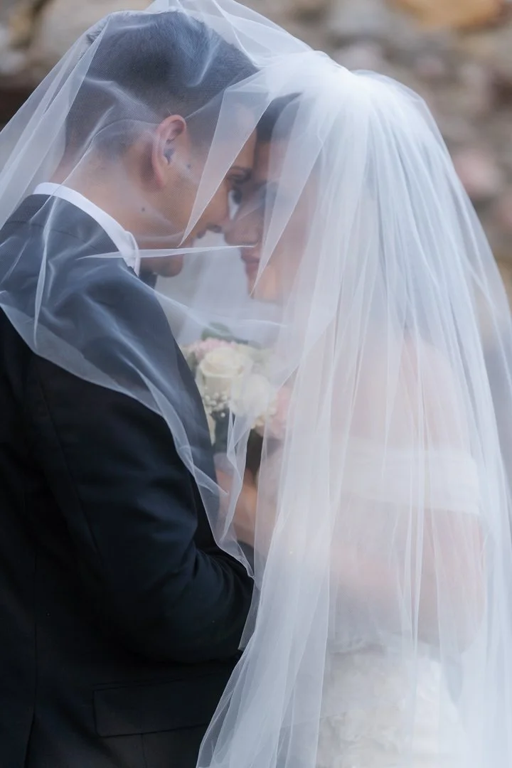 A bride and groom are close together, covered by a sheer wedding veil, with their foreheads touching and smiling at each other during their wedding.