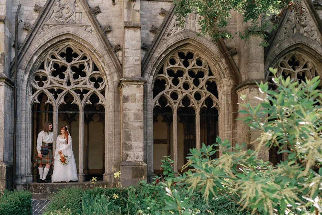 A bride and groom dressed in wedding attire standing together in front of a historic gothic-style stone building with intricate arched windows, surrounded by greenery.
