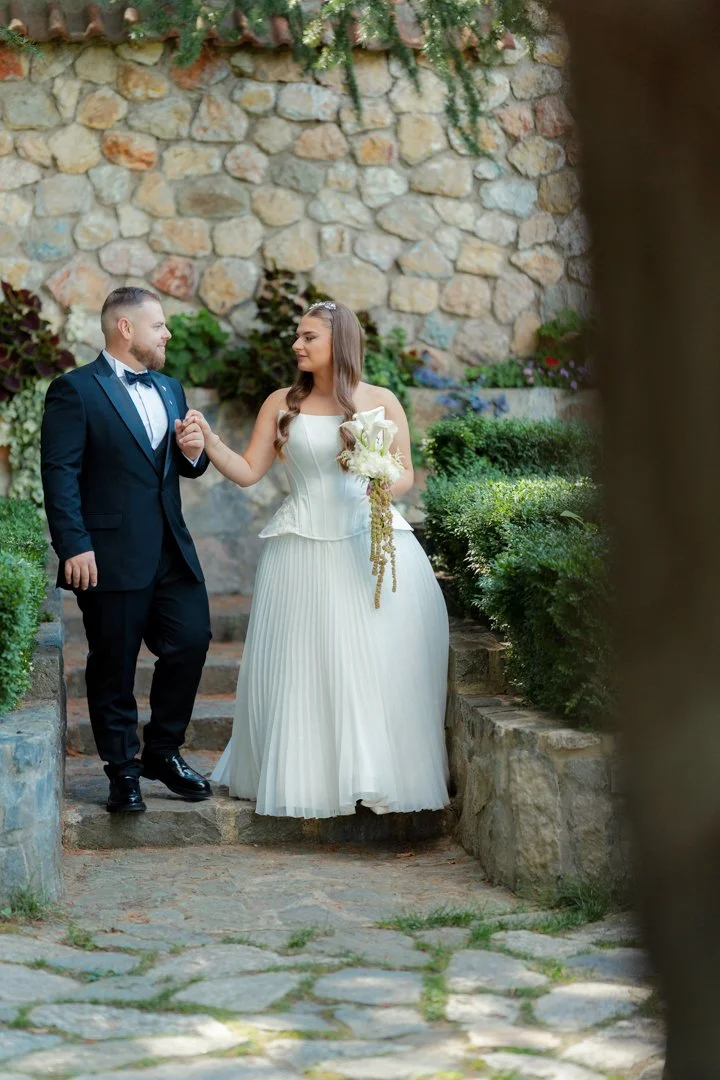 A bride and groom holding hands, walking on stone steps outdoors, with a stone wall and greenery in the background, during a wedding.