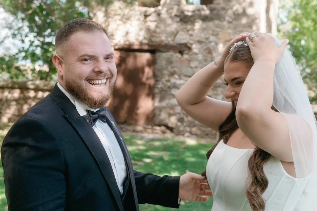 A bride and groom at their wedding outdoors, with the groom smiling and the bride adjusting her veil.