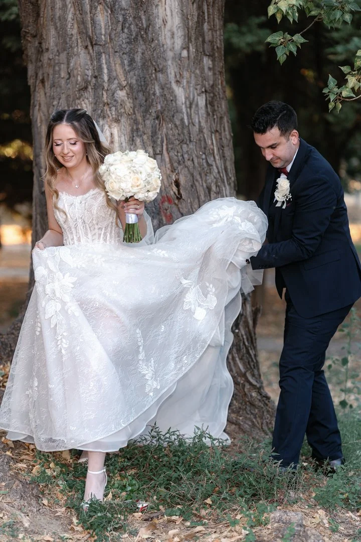 A bride and groom outdoors during their wedding, with the groom lifting the bride's wedding dress while standing near a tree.