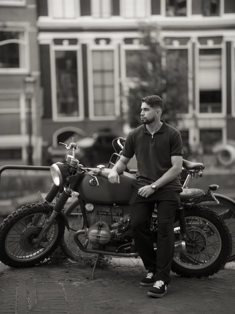 A young man with a beard and short hair sitting on a vintage motorcycle on a city street, wearing a dark polo shirt, dark pants, and sneakers. The background includes buildings with large windows and parked cars, and the image is in black and white.