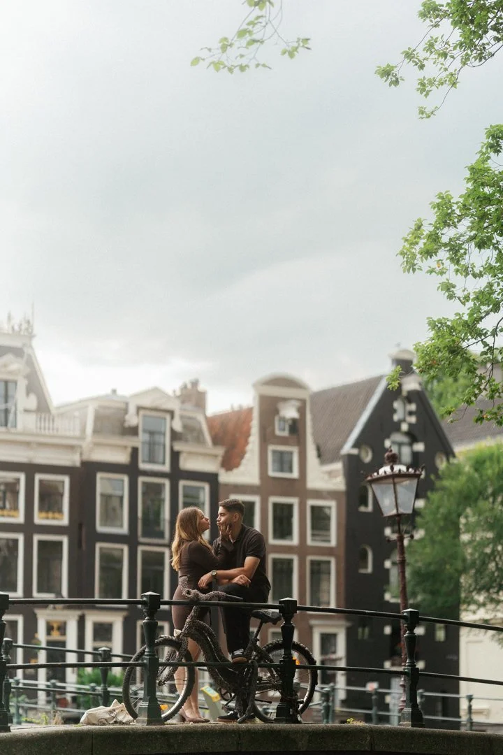 A couple standing close and holding hands on a bridge in front of traditional European style buildings, with one person sitting on a bicycle and they are smiling at each other.
