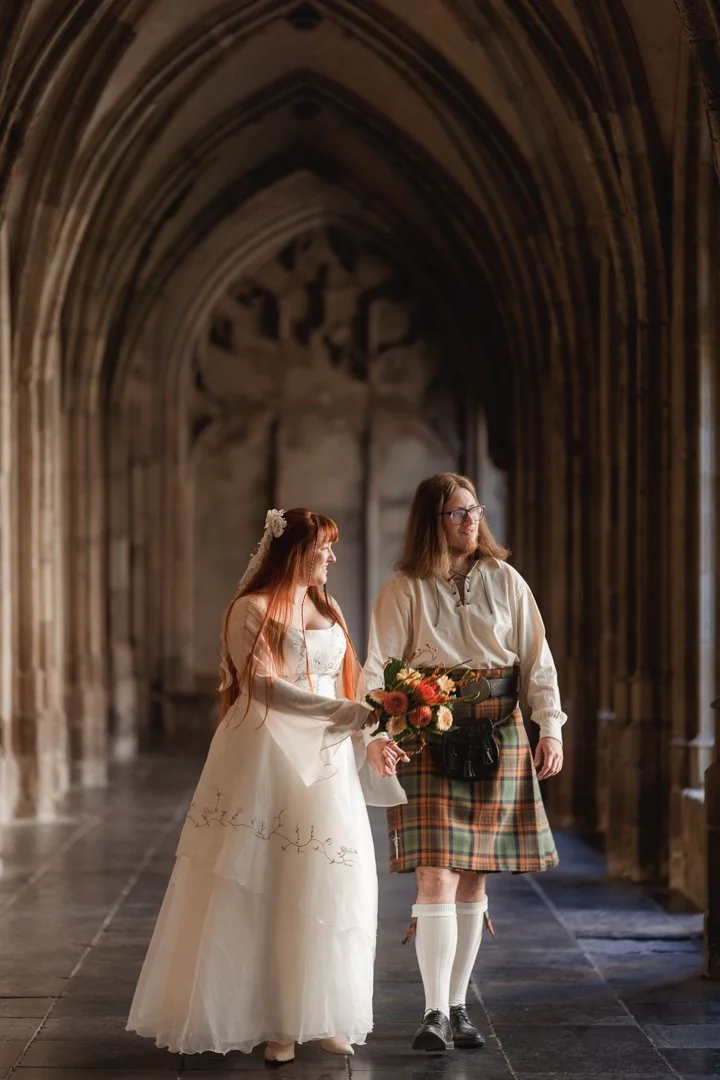 A couple standing in a Gothic cathedral, the woman in a wedding dress holding a bouquet, and the man in traditional Scottish kilt attire, both smiling.