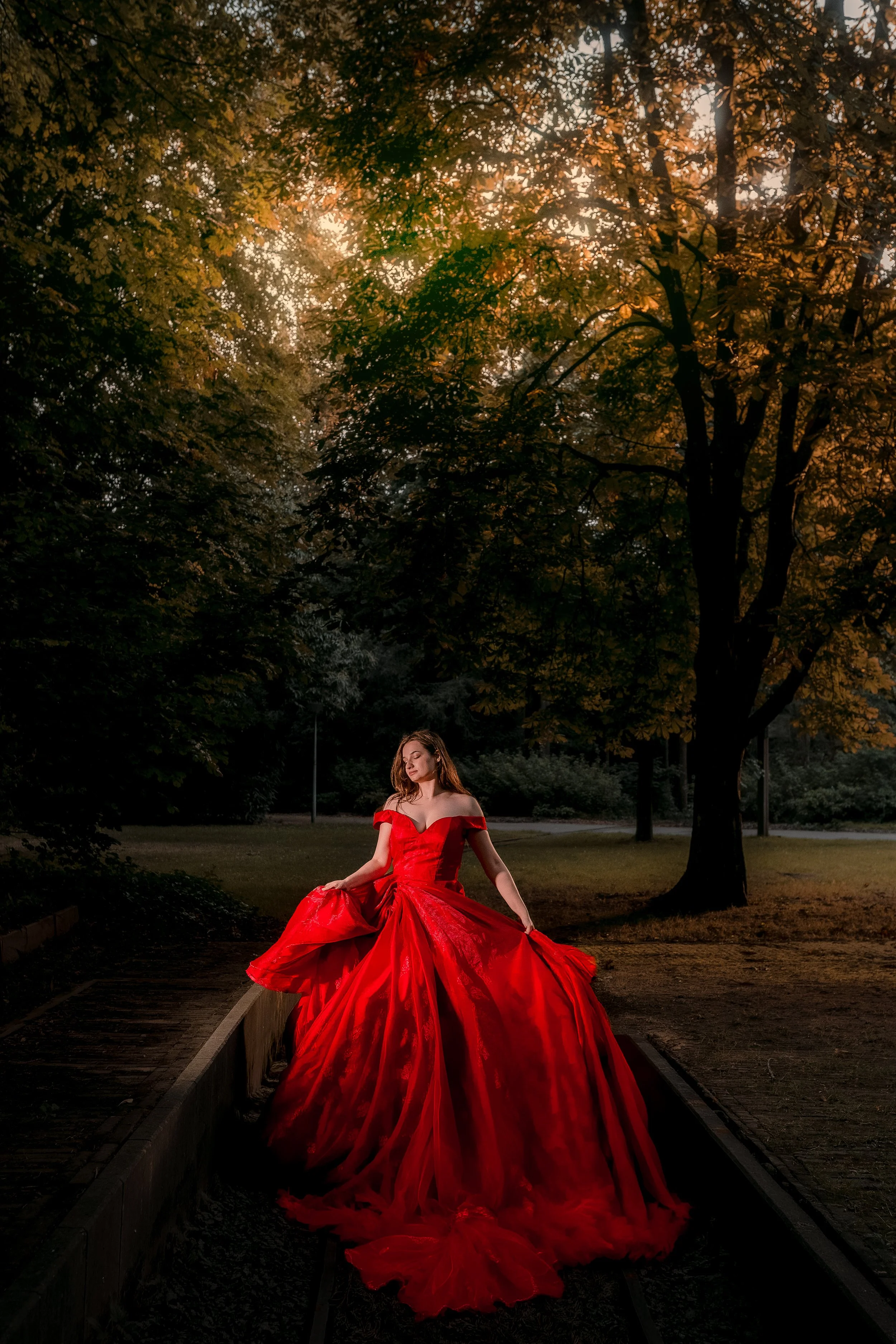 A woman in a flowing, off-the-shoulder, bright red gown sitting on a narrow track in a park at sunset surrounded by trees with golden leaves.