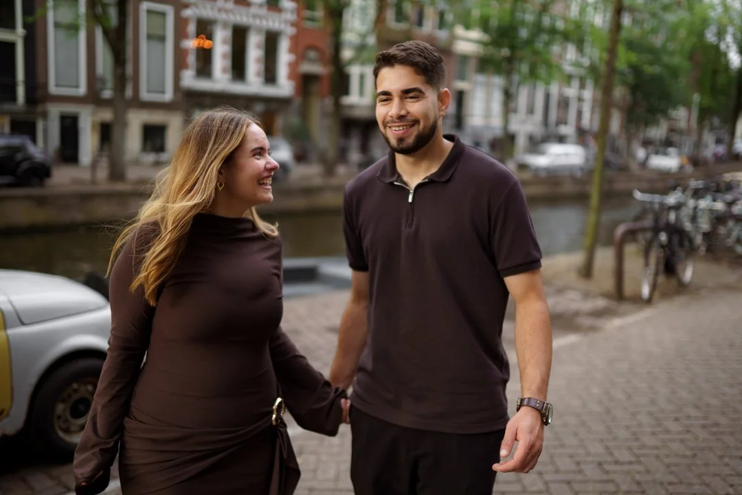 A man and woman holding hands and smiling while walking outdoors near a canal in an urban area, with trees and buildings in the background.
