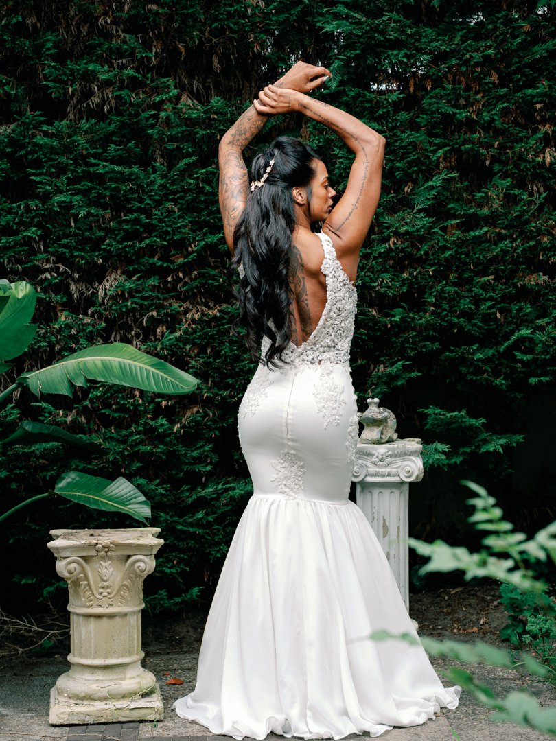 A woman in a detailed white wedding gown with lace and embroidery, posing outdoors with her arms raised above her head, surrounded by greenery and classical stone garden ornaments.