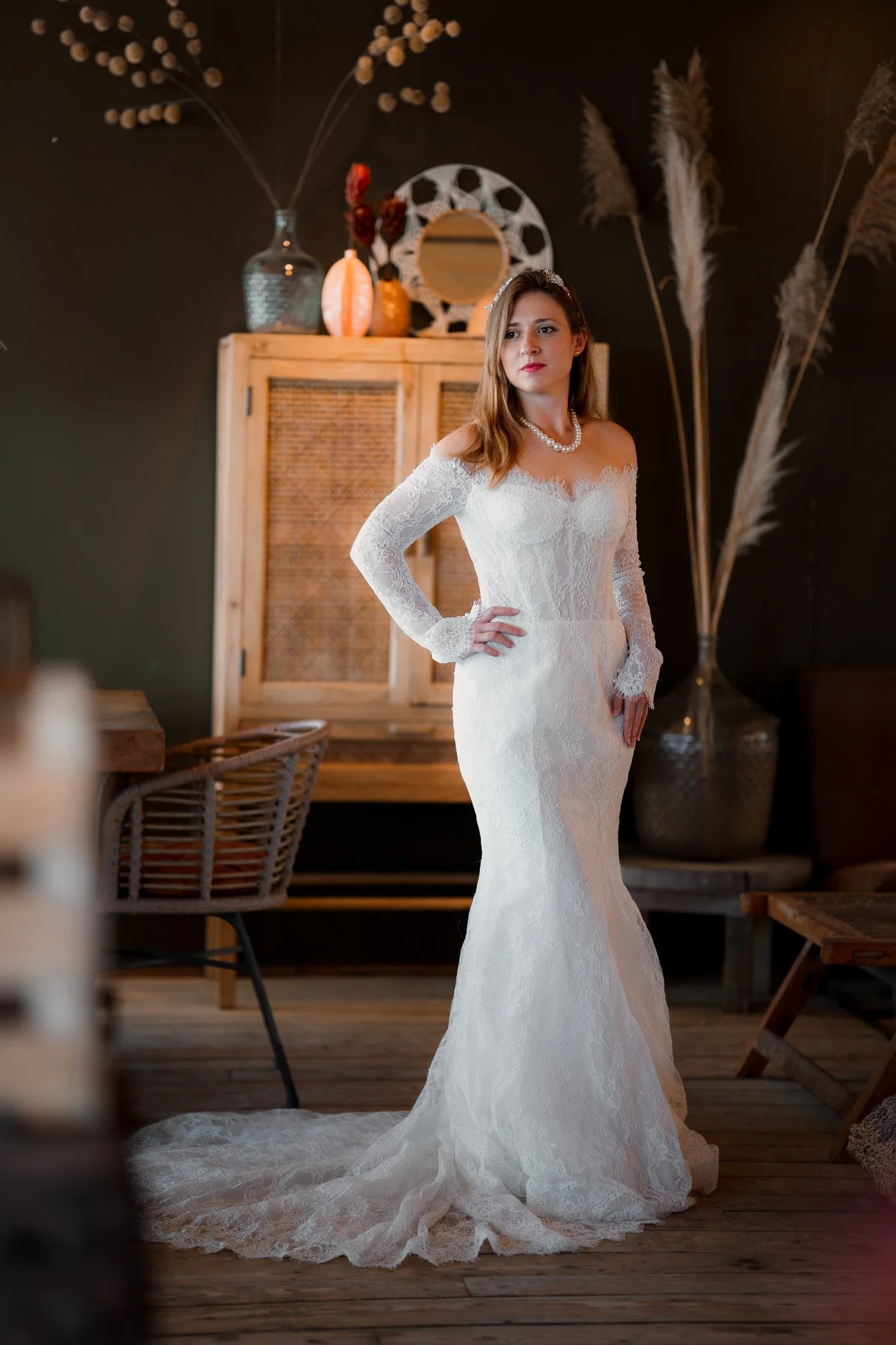 A bride wearing a white lace wedding dress with off-the-shoulder sleeves, a pearl necklace, and a headband, standing in a rustic decor setting with a wooden cabinet, vases, decorative mirror, and tall dried plants.