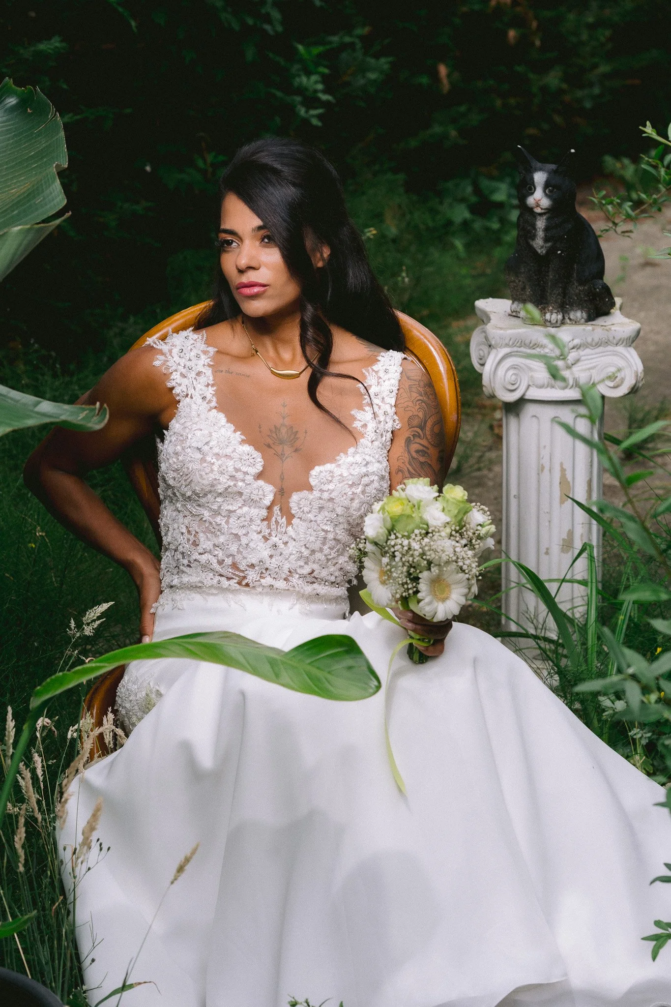 A woman in a white lace wedding dress sitting outdoors surrounded by greenery, holding a bouquet of white flowers, with a black and white cat sitting on a decorative pedestal nearby.