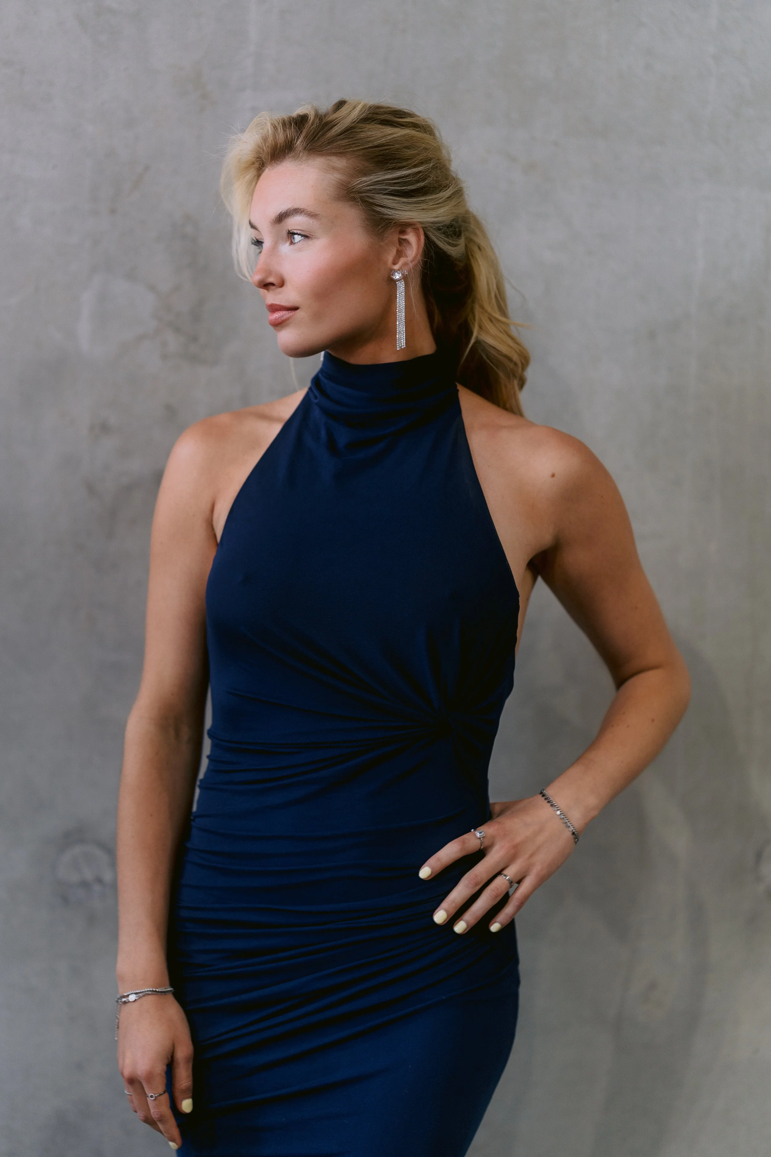 A woman with blonde hair styled in loose waves, wearing a sleeveless dark blue dress with a high neckline, and elegant jewelry including dangling earrings, a bracelet, and rings. She is standing with one hand on her hip against a plain gray background.