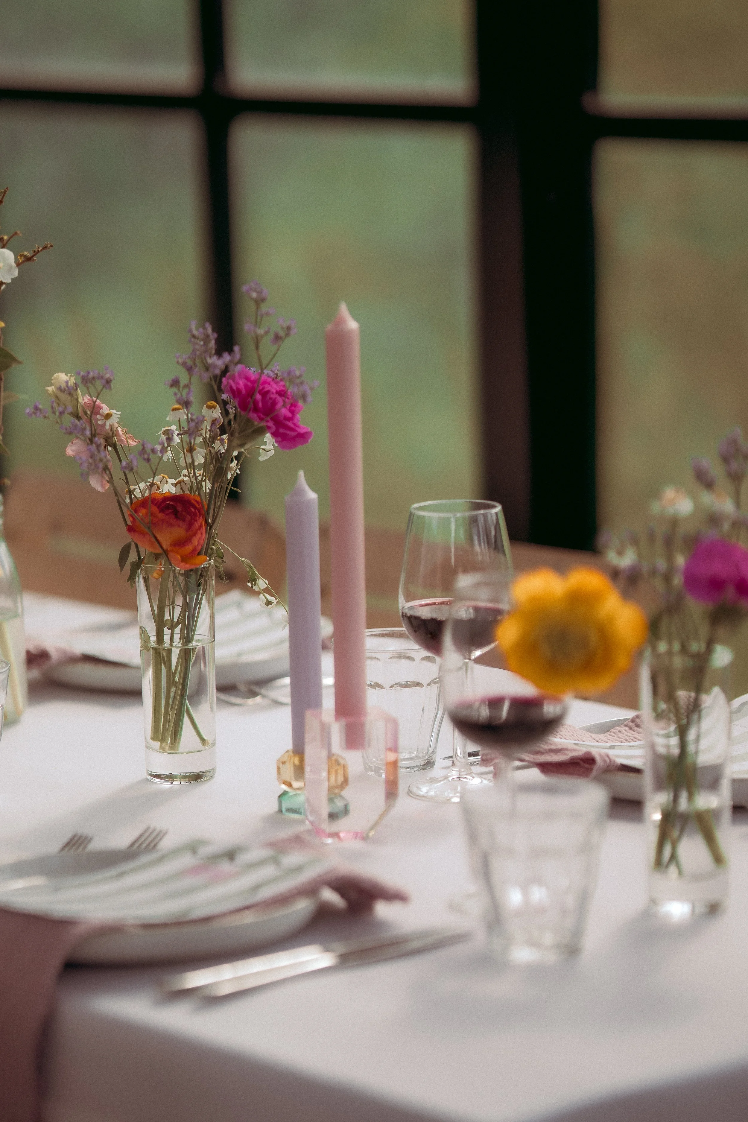 A decorated dining table with a floral centerpiece, pink and purple candles, a wine glass filled with red wine, and tableware set for a meal, with a blurred background of a window or partition.