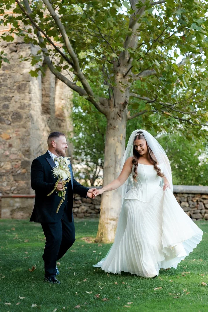 A bride and groom holding hands outdoors on a grassy area with a large tree and stone wall in the background. The bride is wearing a white wedding dress and veil; the groom is in a black suit and bow tie, holding a bouquet.