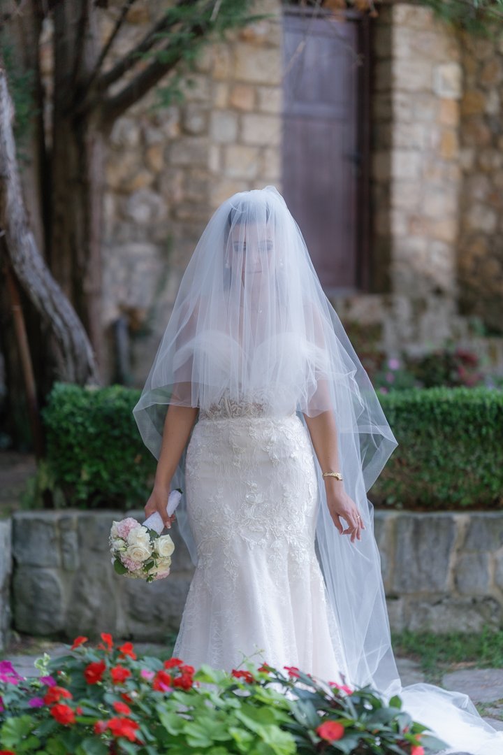 A bride in a wedding dress with a veil standing outdoors, holding a bouquet of flowers, with greenery and stone walls in the background.