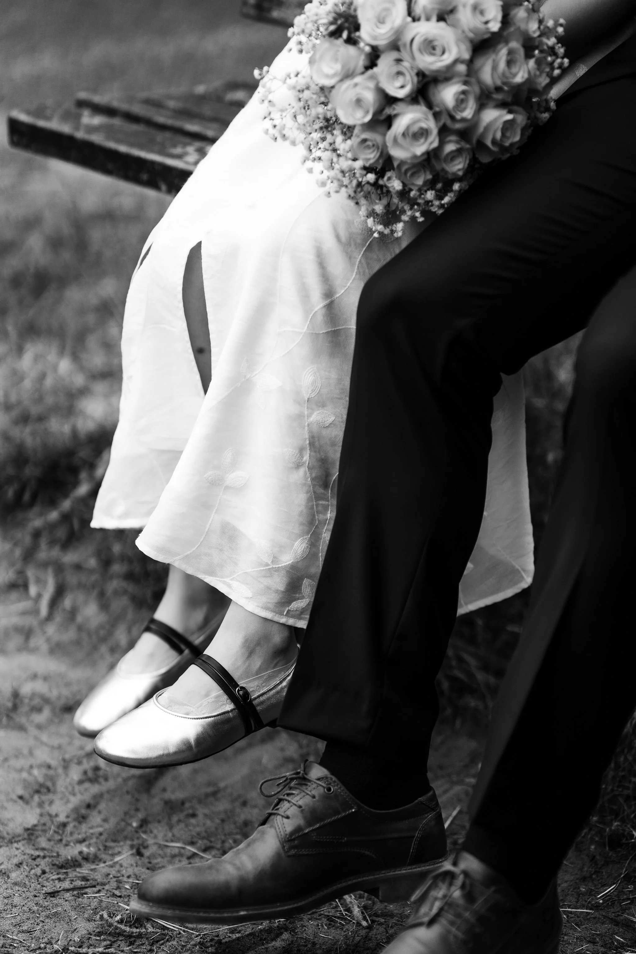 The image shows a pair of wedding shoes worn by a bride, with a bouquet of flowers resting on her lap. The person is sitting on a bench, and only their legs and shoes are visible, with the background blurred.