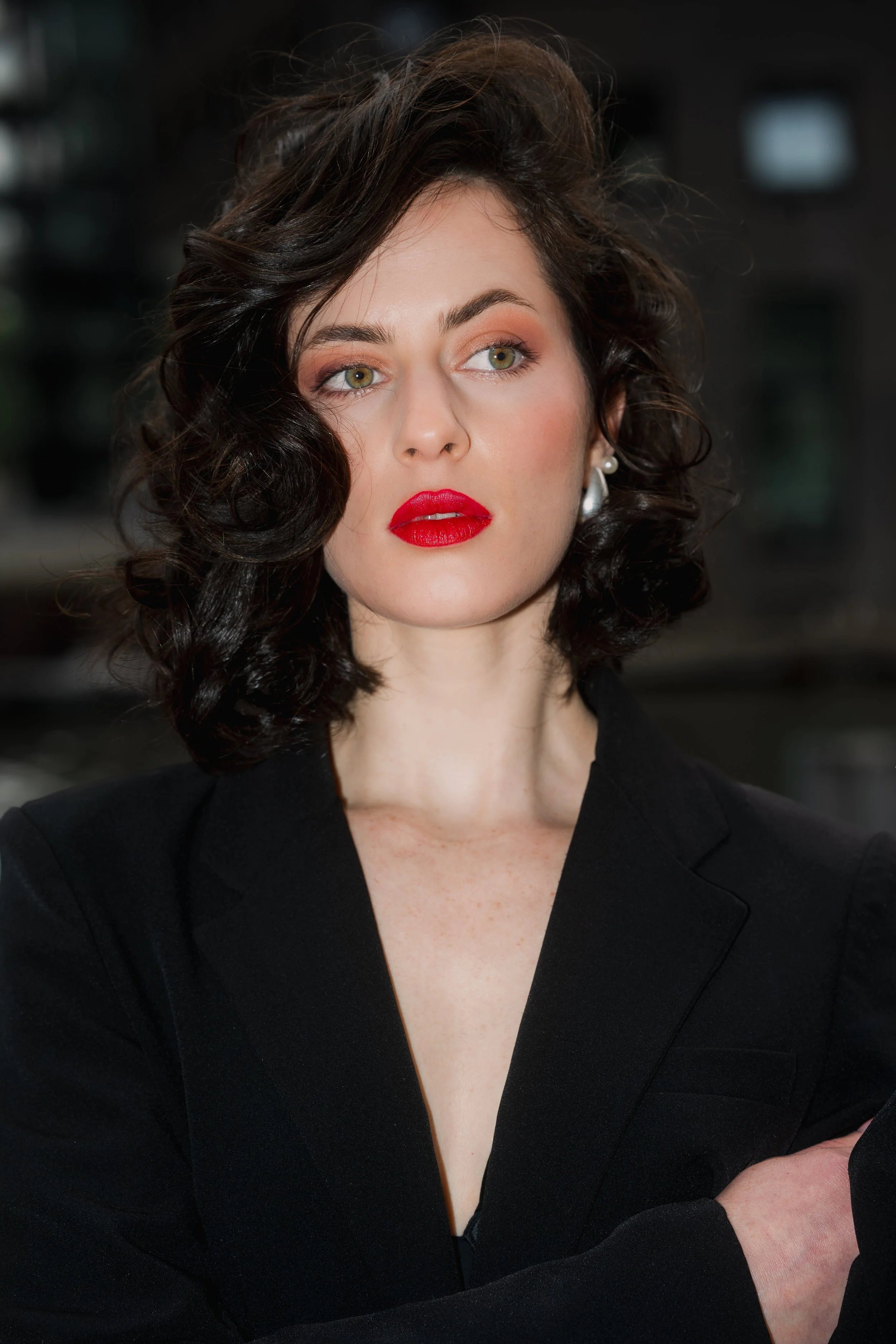 A woman with dark wavy hair, wearing a black blazer, with red lipstick and pearl earrings, looking to the side, standing outdoors.