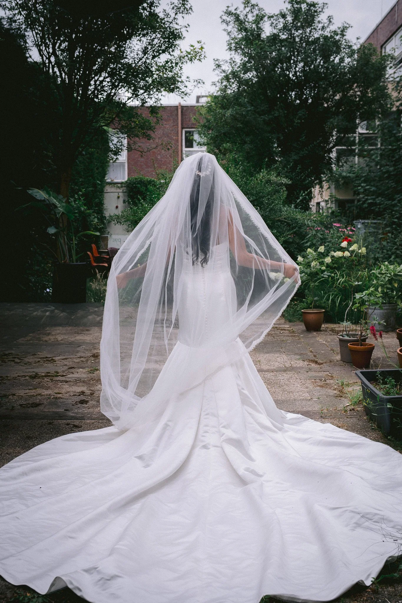 A woman in a white wedding dress and veil standing in a backyard garden, with greenery and potted plants around.