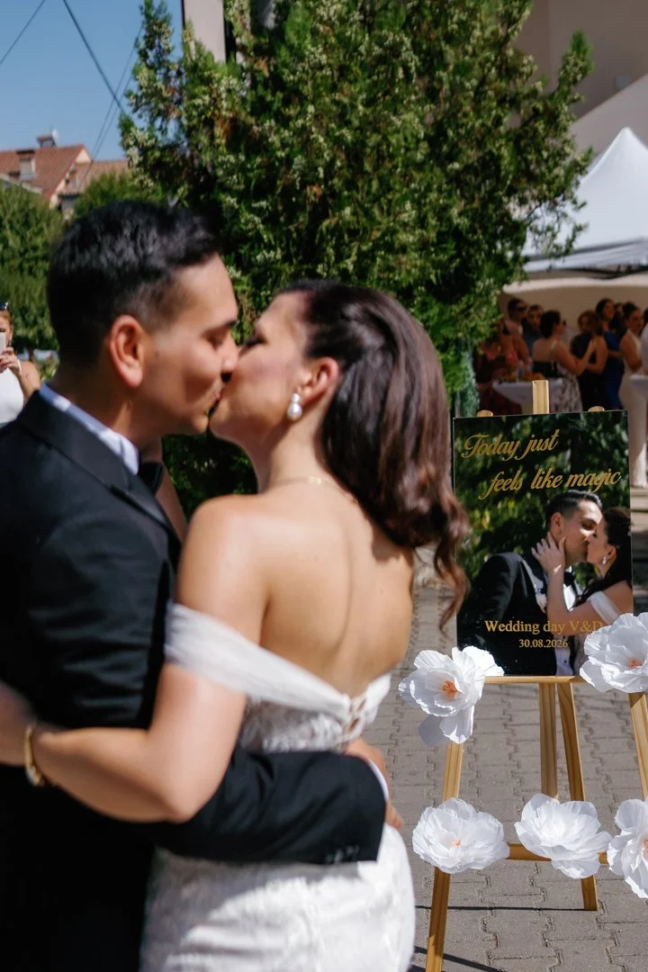A newlywed couple in wedding attire sharing a kiss outdoors. The groom is dressed in a black suit, and the bride is wearing a white strapless wedding dress with pearl earrings. There is a wedding sign on an easel behind them that reads "Today just feels like magic" and includes a photo of the couple along with the date 30.08.2026. The background shows trees, a white tent, and guests at the celebration.