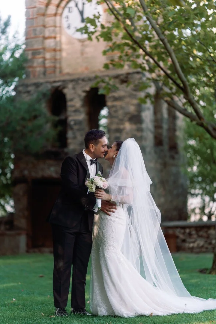 Bride and groom in wedding attire sharing a kiss in a park with an old stone clock tower in the background.