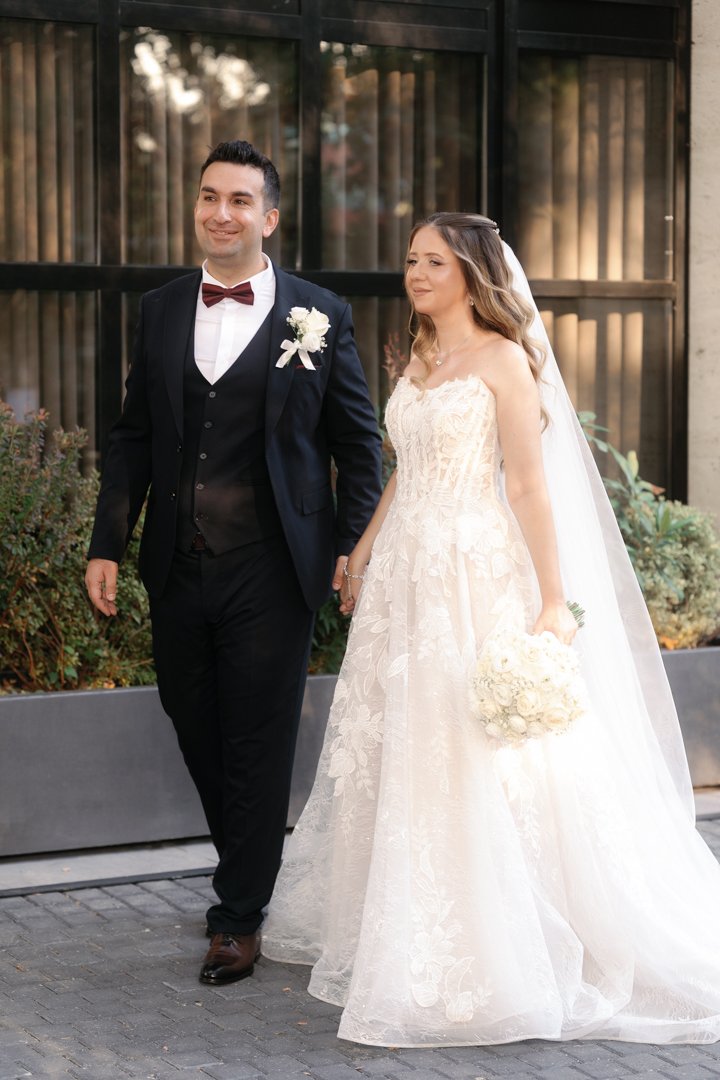 A bride and groom walking hand in hand outdoors, both smiling, in front of a modern building with large windows. The bride is wearing a strapless white wedding gown with lace details and a veil, holding a bouquet of white flowers. The groom is wearing a black suit with a white shirt, a red bow tie, and a white boutonniere.