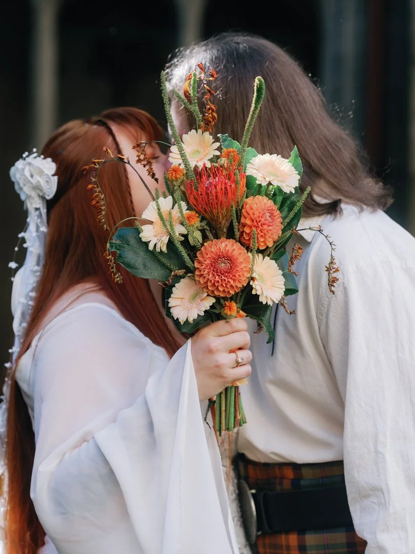 Two people are kissing behind a large bouquet of flowers, with only parts of their faces visible.
