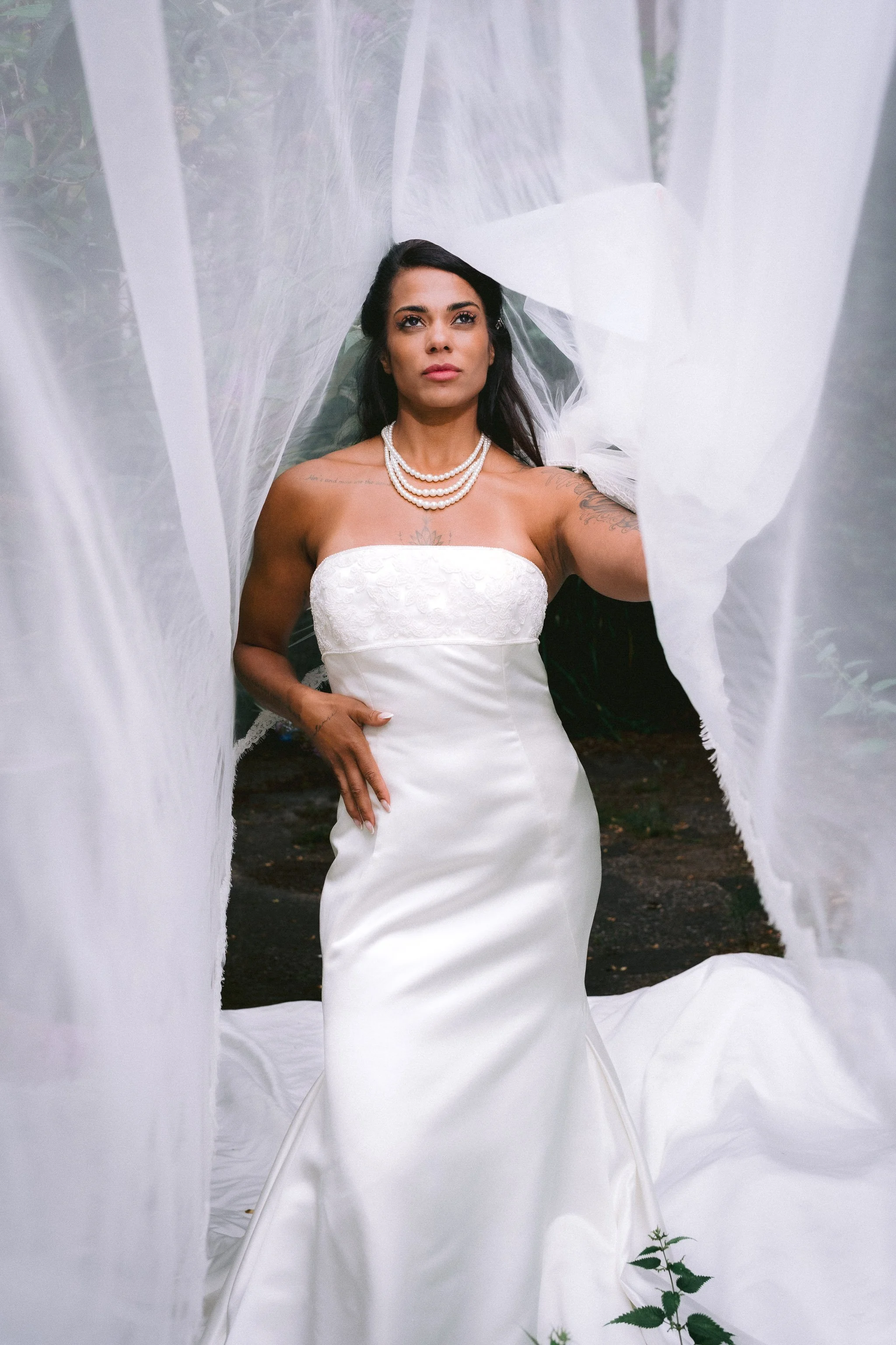 A woman in a white strapless wedding dress and pearl necklace standing outside among white fabric drapes, with plants visible in the background.