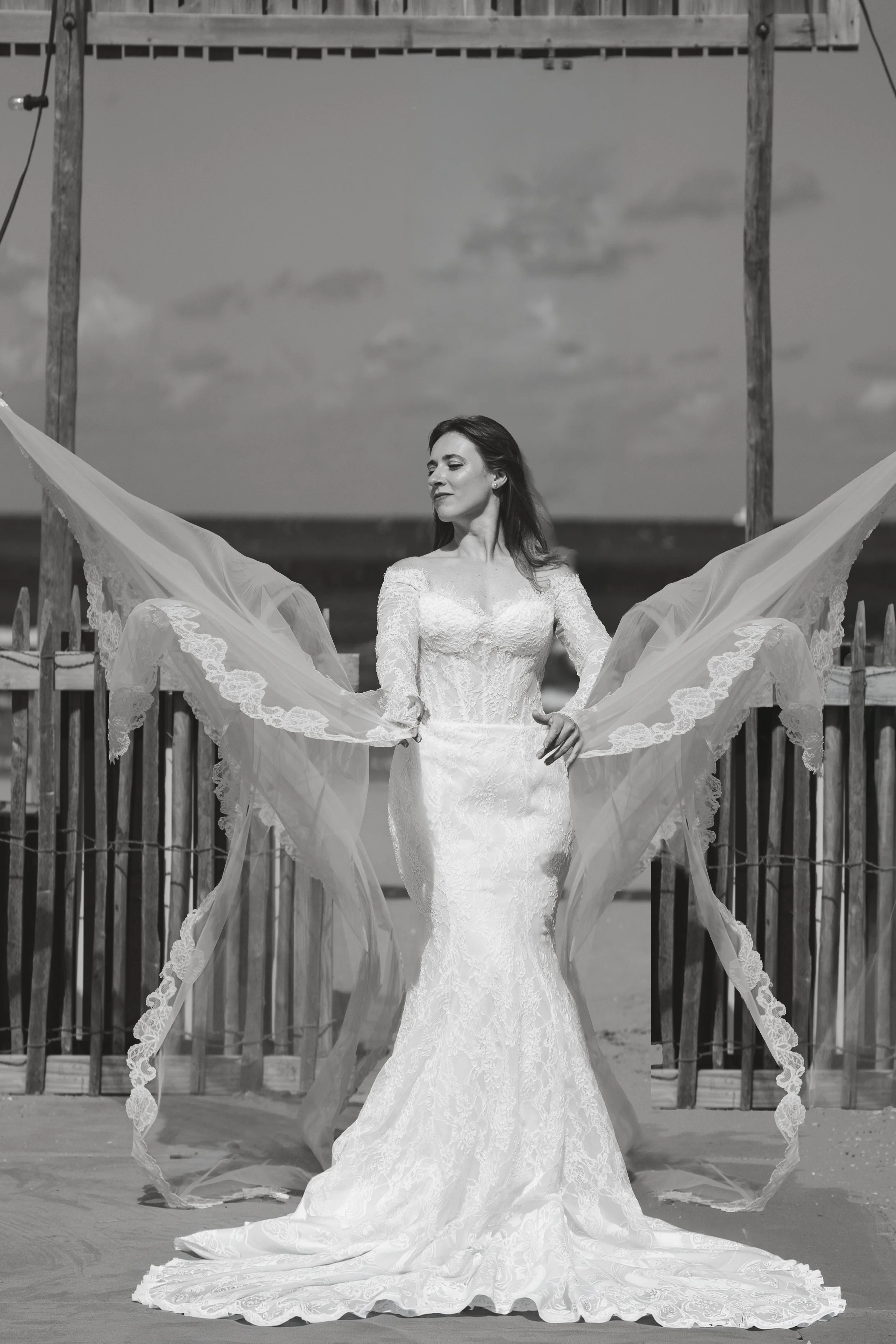 A bride in a long, elegant lace wedding dress standing outdoors on a wooden deck, holding up the sides of her lace veil with both hands, with a wooden fence, cloudy sky, and ocean in the background.
