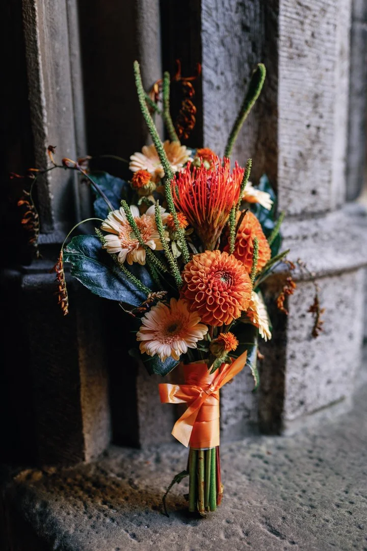 A bouquet of orange and peach flowers, tied with an orange ribbon, placed against a stone wall.