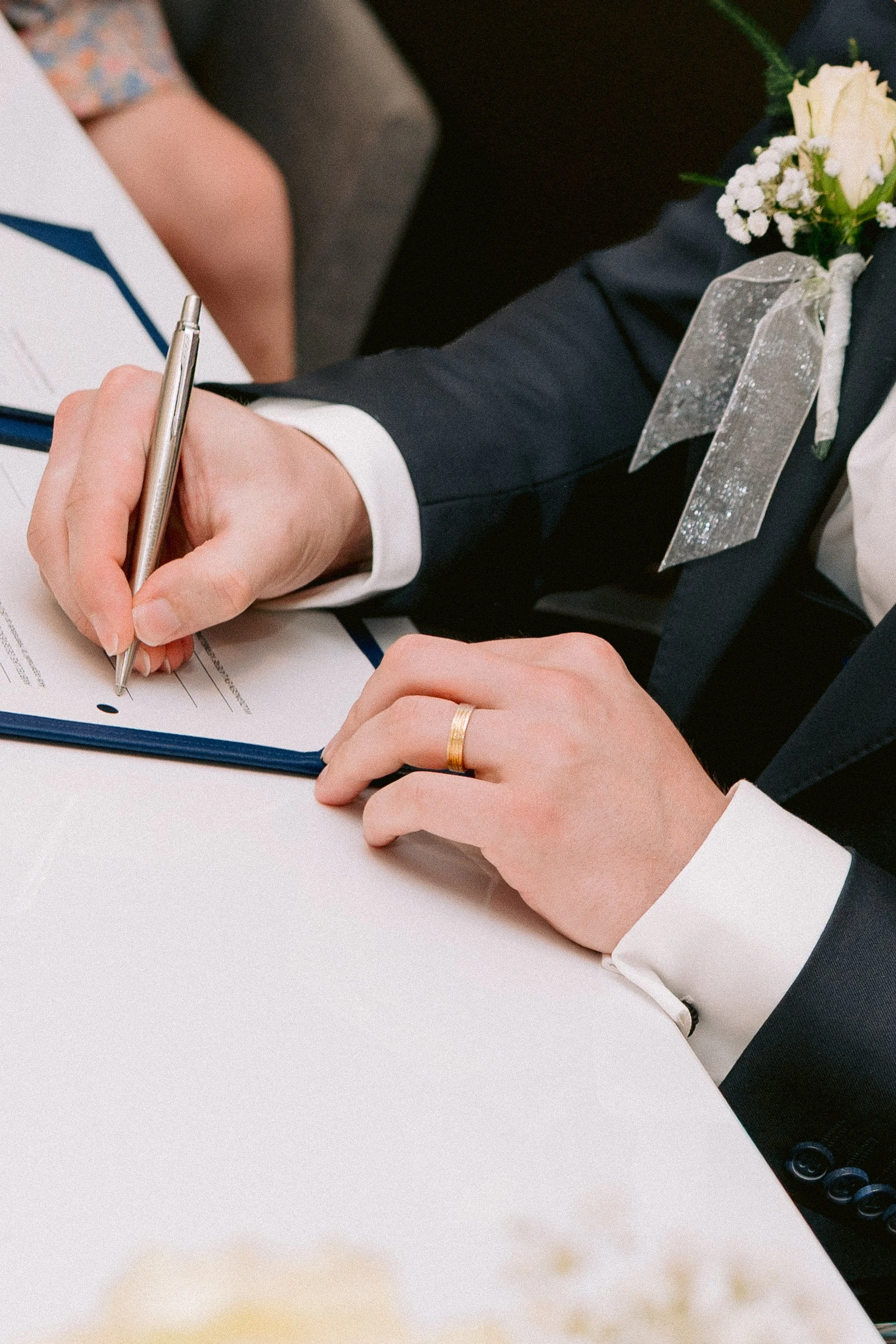 A person wearing a dark suit with a white shirt is signing a document with a silver pen. They have a ring on their finger and a corsage pinned to their suit jacket. The setting appears to be a formal event or ceremony.