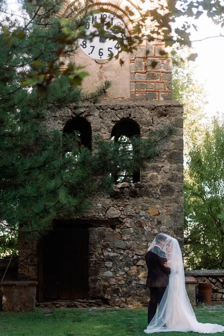 A bride and groom standing close under a wedding veil outdoors near a stone tower with a clock, surrounded by trees.