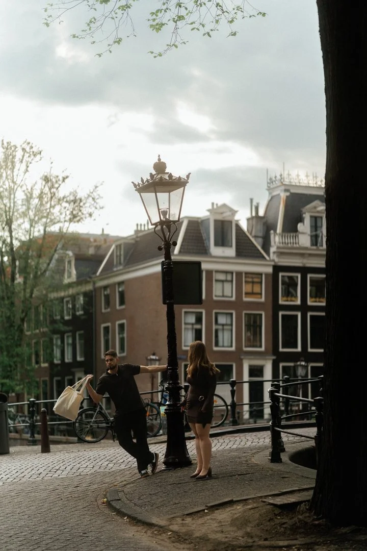 A man and woman standing near a streetlamp on a city sidewalk with traditional European-style buildings in the background. The man appears to be talking and holding a bag, while the woman listens.