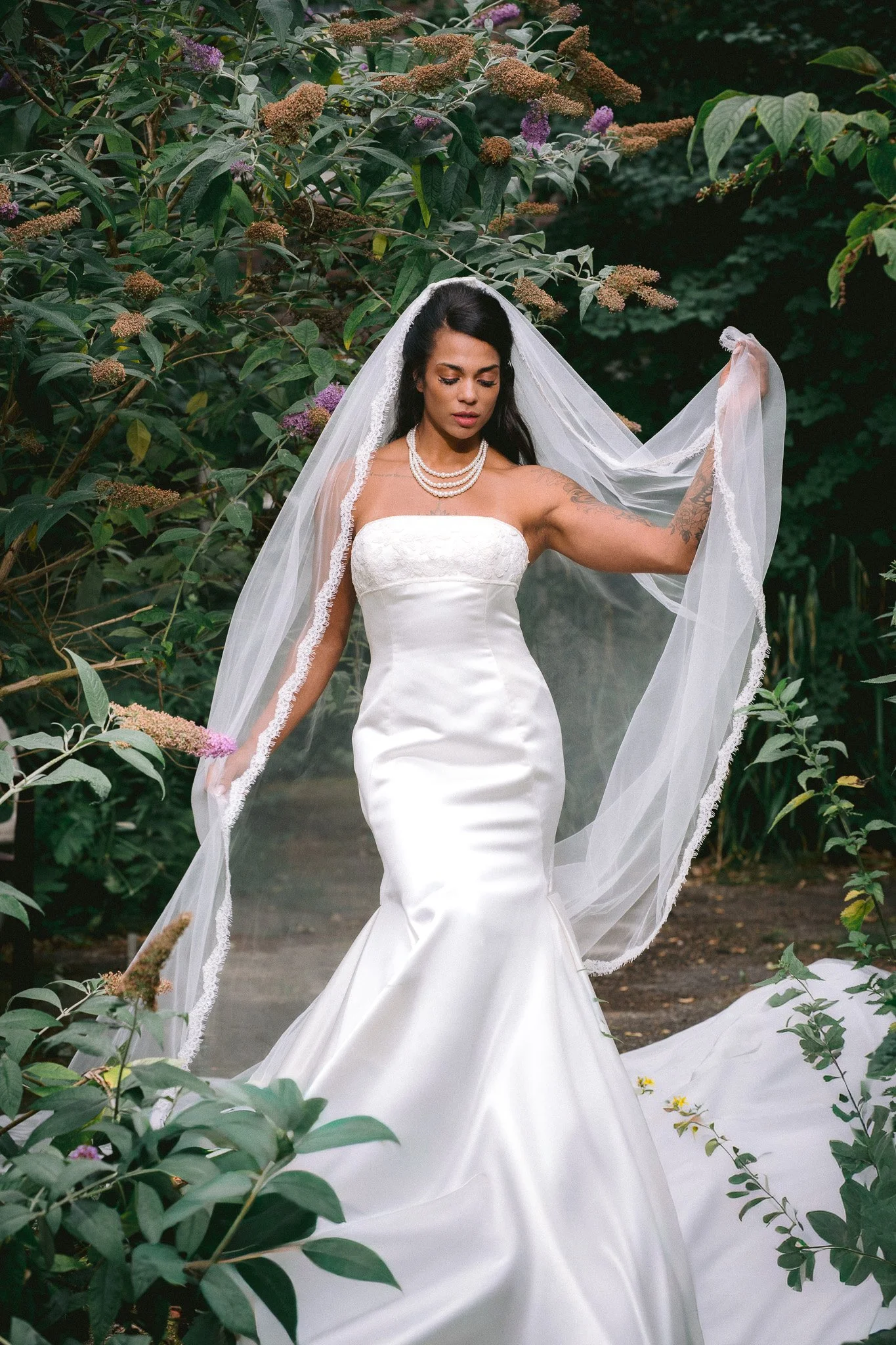 A woman dressed in a wedding gown and veil stands outdoors among green bushes with purple and pink flowers, holding the edge of her veil.